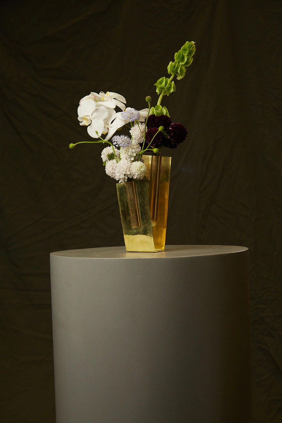 Vaso-escultura Vértice do Ouro em latão, com flores brancas, sobre pedestal cinza